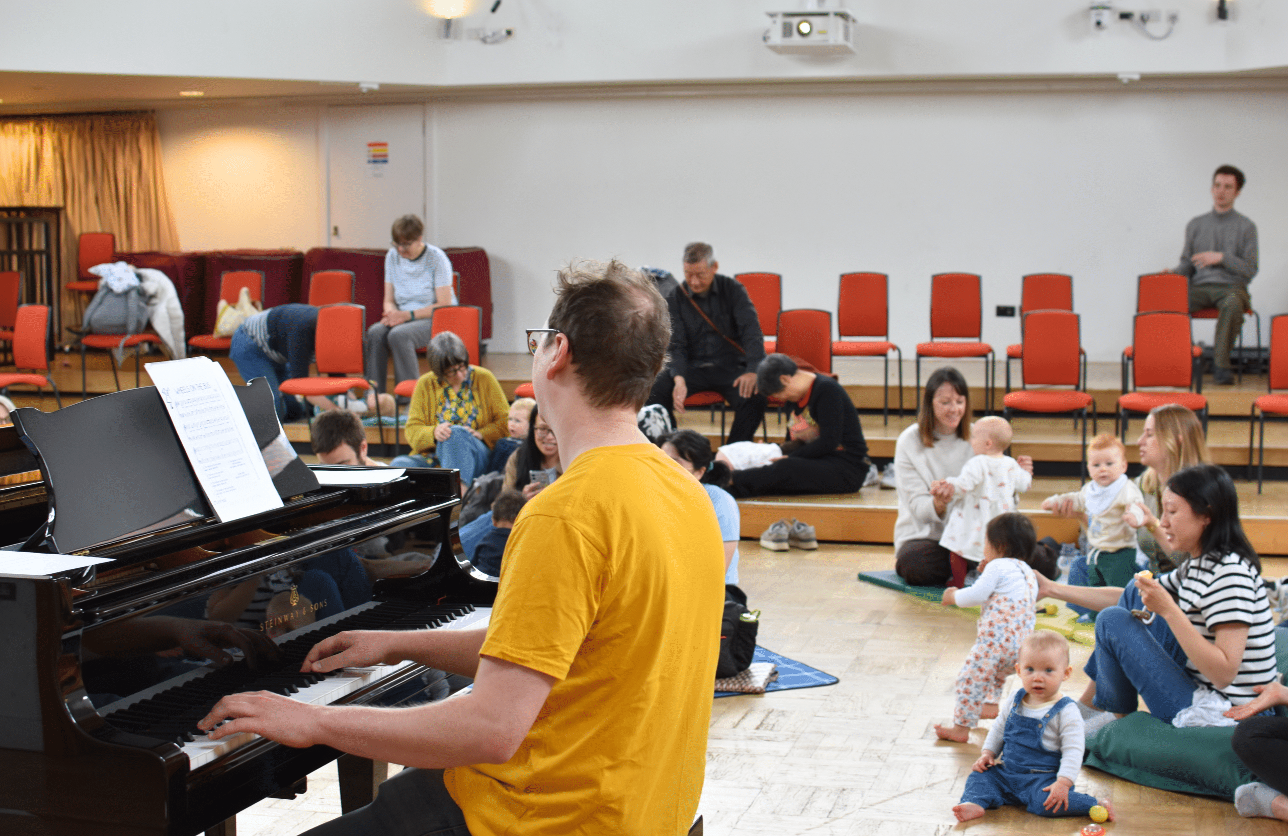 A man in yellow plays piano while babies dance on mats and beanbags