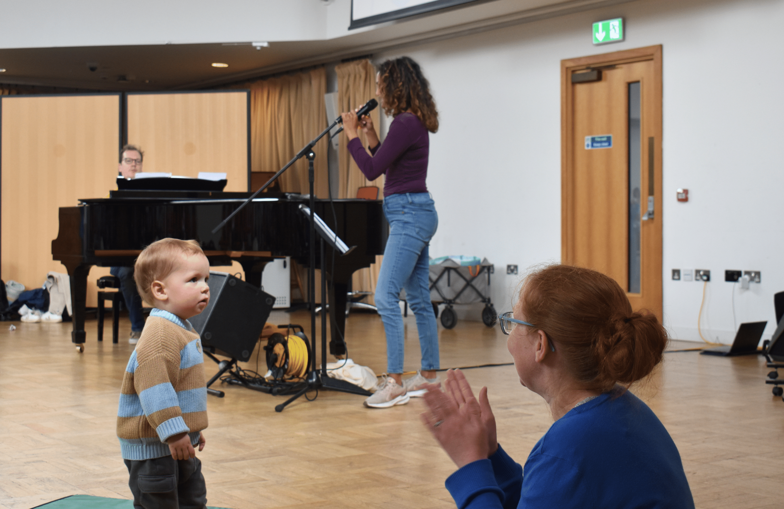 A small child looks at a woman clapping. A singer and piano player are behind.